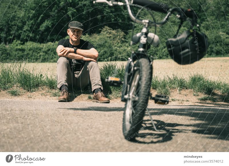 Mann sitzt hinter einem Kinderfahrrad im Gras Fahrrad Fahrradfahren Fahrradhelm Kinderrad sitzen Sport Pause Natur Außenaufnahme Freizeit & Hobby Farbfoto