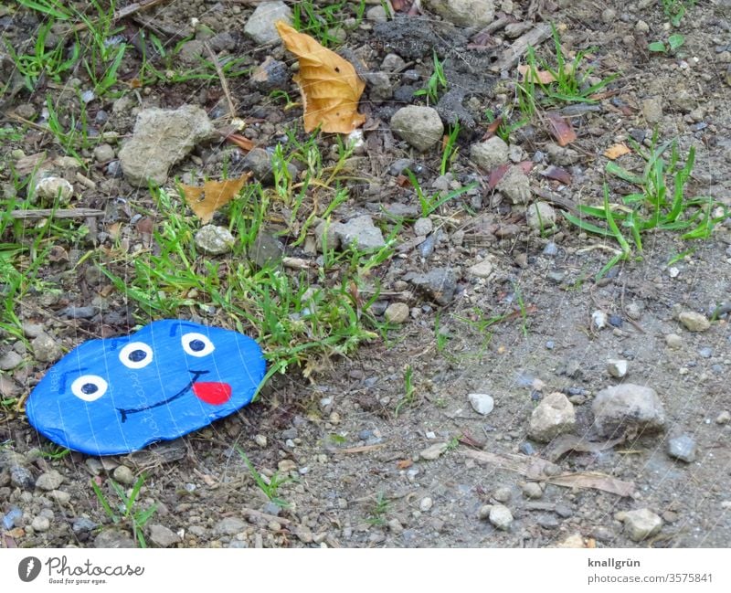 Ein blauer Glücksstein mit einem lustigen Monstergesicht mit drei Augen und herausgestreckter Zunge am Wegesrand Glückssteine Freude Wunschstein bemalt