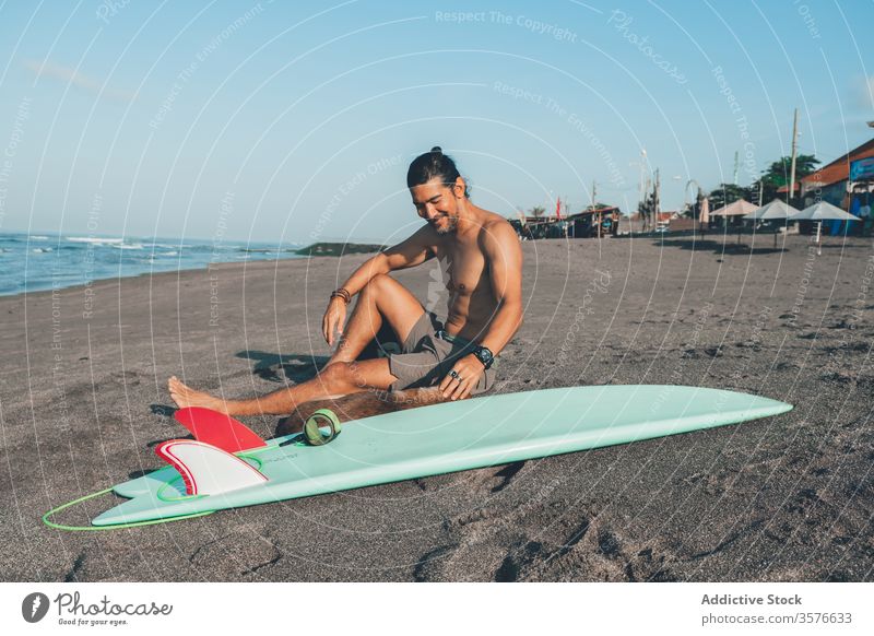 Männlicher Surfer mit Hund, der sich am Strand ausruht Mann ruhen Zusammensein Haustier Sand sitzen Freund Surfbrett Begleiter jung ohne Hemd männlich Sommer