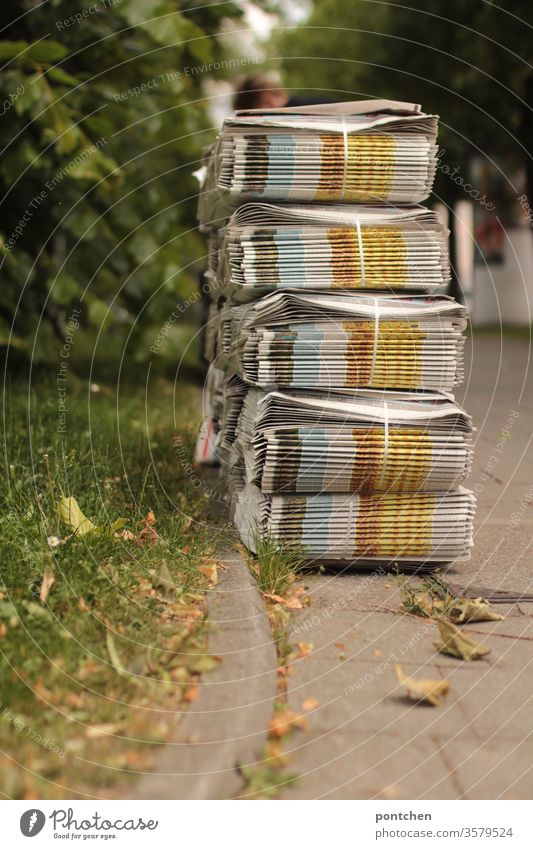 Viele Stapel an Zeitungen Wochenblättern stehen auf dem Gehweg und warten auf Abholung durch den Zeitungsausträger. Eine Frau steht gebückt über ihnen