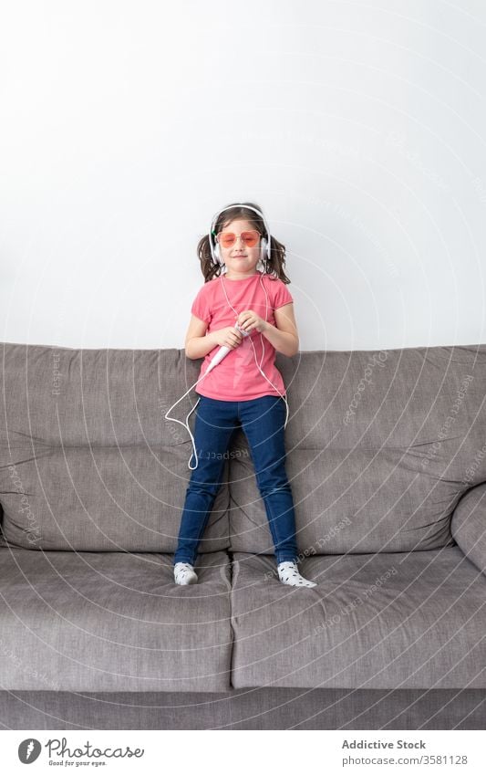 Glückliches Mädchen im Vorschulalter mit Kopfhörern und Mikrofon singt, während sie alleine zu Hause spielt singen Spaß haben Kind Sofa heimwärts Sänger spielen