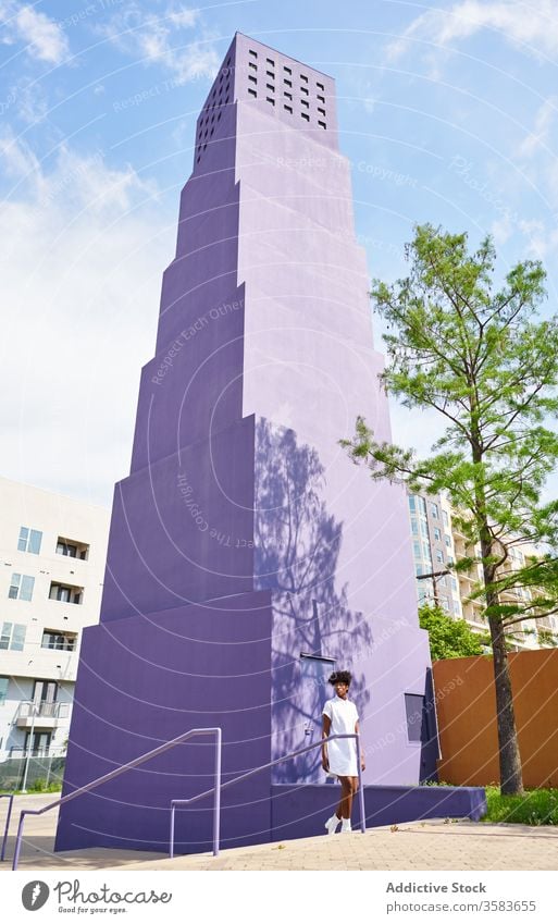 Elegante schwarze Frau geht auf der Straße gegen ein lilafarbenes Hochhaus in der Innenstadt laufen Kleid Mode trendy Outfit Architektur Stil hoher Anstieg