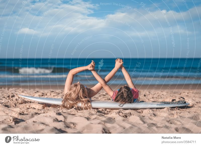 Zwei kleine Mädchen halten die Hände zusammen, die auf einem Surfbrett am Sandstrand des Ozeans liegen. Liebe, Freundschaft, Zusammengehörigkeitskonzept.