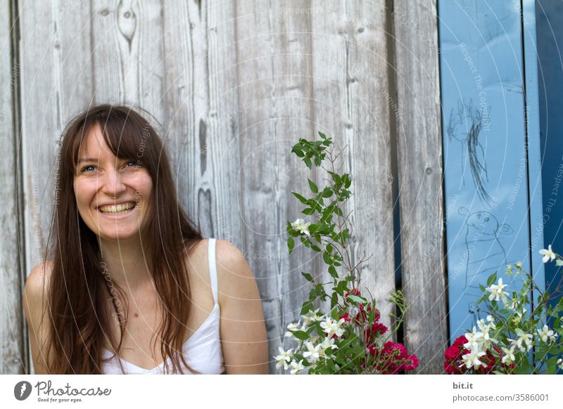 Lachende, langhaarige junge Frau sitzt vor einer alten Holzwand draussen im Garten und freut sich über Blumen-Geschenk von Partygast. Fröhliche, natürliche, brünette junge Erwachsene mit strahlendem Gesicht auf Gartenparty Fest in Partylaune lacht freudig.