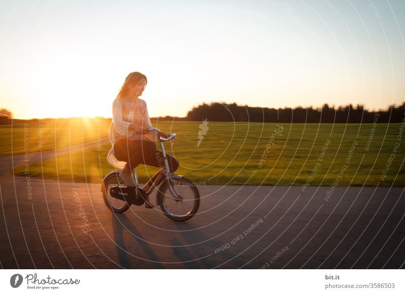 Langhaarige, brünette Jugendliche, fährt bei Abendlicht mit einem kleinen Fahrrad auf der Straße. Fahrradtour in der Dämmerung, vorbei an grüner Wiese und Wald, wobei der Teenager, wie ein lustiger Riese auf dem überdimensional, winzigen Zwergen-Rad wirkt.