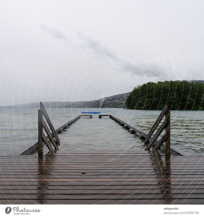 Nasse Holzbretter eines Badestegs, ein Holzgeländer in den See, eine Absperrung im See, eine blaue Badeinsel, alles bei regnerisch grauem Wetter Badeanstalt