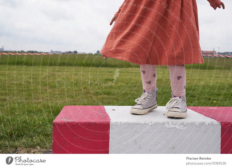 Mädchen mit Kleid und Turnschuhen steht auf einer rot weiß gestreiften Betonabsperrung auf dem Tempelhofer Feld, im Hintergrund eine Wiese und rot weißes Absperrband