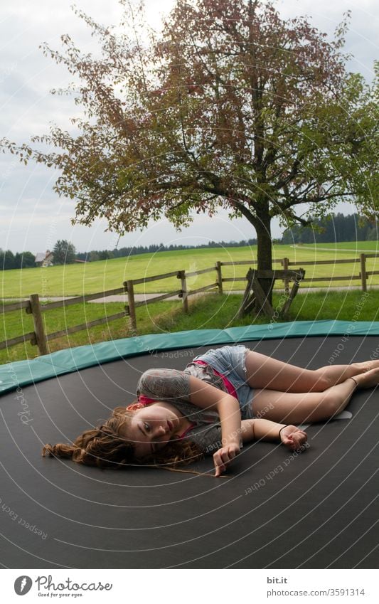 Müder, weiblicher Teenager liegt faul auf Trampolin im Garten und betrachtet seine lockigen Haare. Langhaarige, brünette Jugendliche chillt auf Trampolin in der Natur und sucht Haarspliss in Haarsträhne. Pause, Ausruhen, Entspannen, Erholen nach Sport.