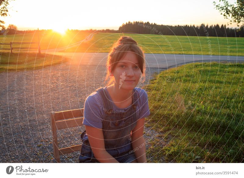 Teenager sitzt bei Sonnenuntergang auf einem Weg mit Kieselsteinen, auf einem Holzstuhl und schaut in die Kamera. Jugendliche genießt sitzend die Abendsonne, im Sommerurlaub auf dem Land, vor grüner Landschaft, Wiesen, Weide, Wald und warmen Sonnenlicht.