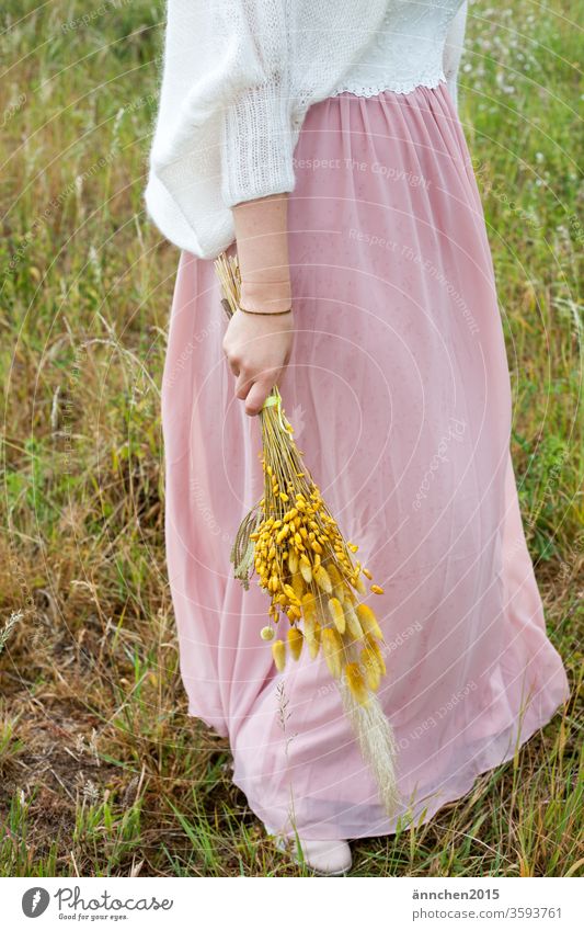 Frau hat einen rosa Rock und ein weißes Oberteil an, über dem Oberteil ist eine weiße leichte Jacke sie hält Blumen in der Hand kleid Blumenstrauß Trockenblumen