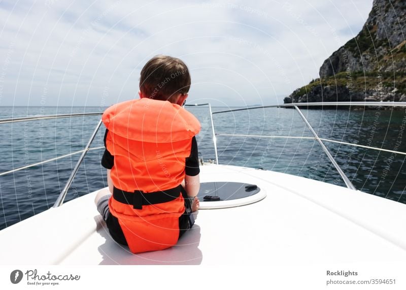 Rückansicht eines Jungen, der auf dem Deck eines Motorbootes sitzt und eine orangefarbene Schwimmweste trägt kleiner Junge Kind Sitzen Schiffsdeck Boot