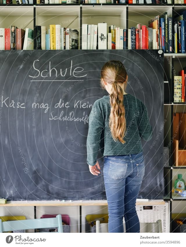 Homeschooling I - Mädchen schreibt mit Kreide auf eine Tafel einen Übungssatz für Schreibschrift, im Hintergrund Bücherregal Schule Schülerin Grundschule Regal