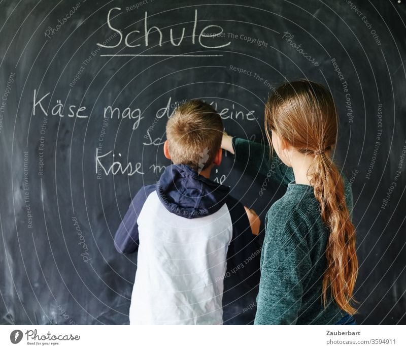 Homeschooling IV - Kinder spielen Schule, eines schreibt mit Kreide auf eine Tafel einen Übungssatz für Schreibschrift Junge Schülerin Grundschule Bücher Regal