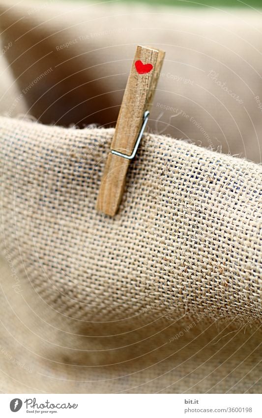 Alte, braune Wäscheklammer aus Holz mit kleinem, rotem Herz klammert an einem Stoffstück aus Jute. Symbol für Nachhaltigkeit durch abbaubare Verpackungen und EInkaufen mit Taschen aus natürlichem Material. Zeichen für Liebe, Freundschaft und Zusammenhalt.