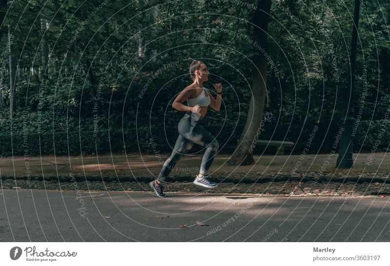 Selektiver Fokus in der Bewegung einer hübschen Sportlerin, die an einem Sommernachmittag eine von Bäumen umgebene Straße hinunterläuft. Fitte Frau beim körperlichen Training.