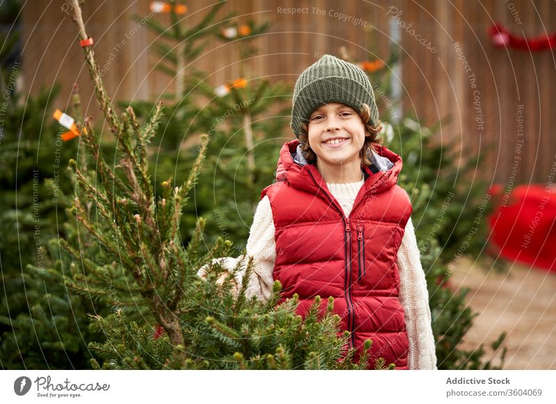 Schöner blonder Junge mit grüner Wollmütze, roter Weste, weißem Pullover, blauer Hose und gelben Stiefeln wählt seinen Weihnachtsbaum frisch Nahaufnahme Jungen