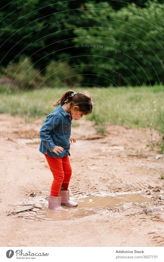 Neugieriges Mädchen mit chirurgischer Maske spielt in Schlammpfütze Kind Pfütze Spaß haben neugierig Gummi Stiefel niedlich dreckig Wasser nass bezaubernd