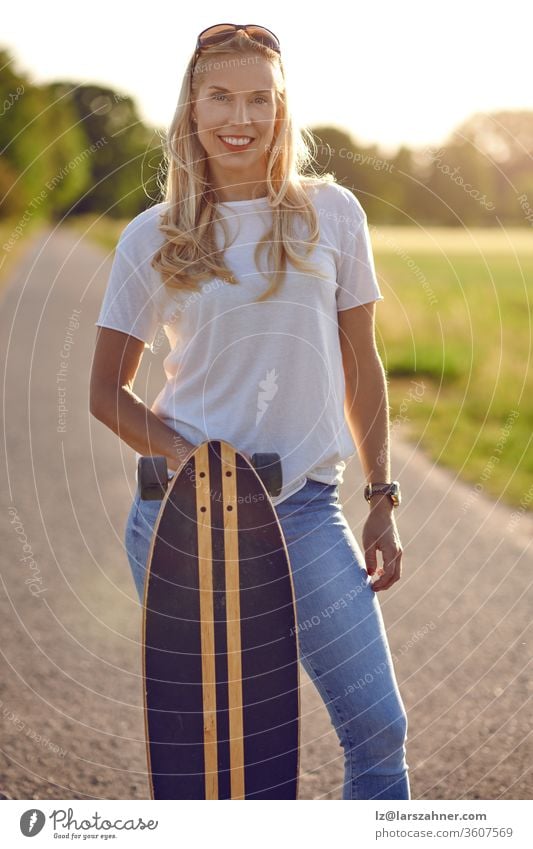 Porträt einer fitten schönen Frau mittleren Alters mit aktivem Lebensstil, die lächelt und in die Kamera schaut, während sie im Sommer auf einer sonnigen Straße im Park ein Longboard hält