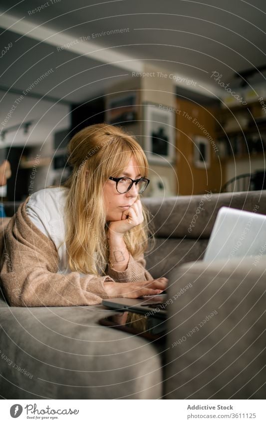 Nachdenkliche Frau arbeitet zu Hause am Laptop Browsen freiberuflich Projekt benutzend Telearbeit Lügen Sofa Arbeit abgelegen ernst Brille Apparatur Internet