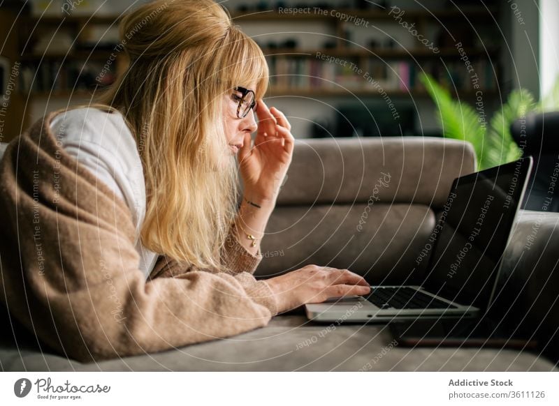 Nachdenkliche Frau arbeitet zu Hause am Laptop Browsen freiberuflich Projekt benutzend Telearbeit Lügen Sofa Arbeit abgelegen ernst Brille Apparatur Internet