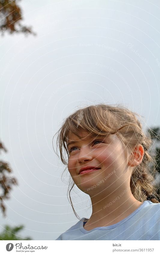 Blondes, natürliches, fröhliches Mädchen steht lächelnd und zufrieden, draussen in der Natur, zwischen grünen Bäumen, vor bedecktem Himmel. Glückliches, strahlendes Mädchen, bei Familienausflug im Frühling, mit wehenden Haaren in der hellen Umgebung..