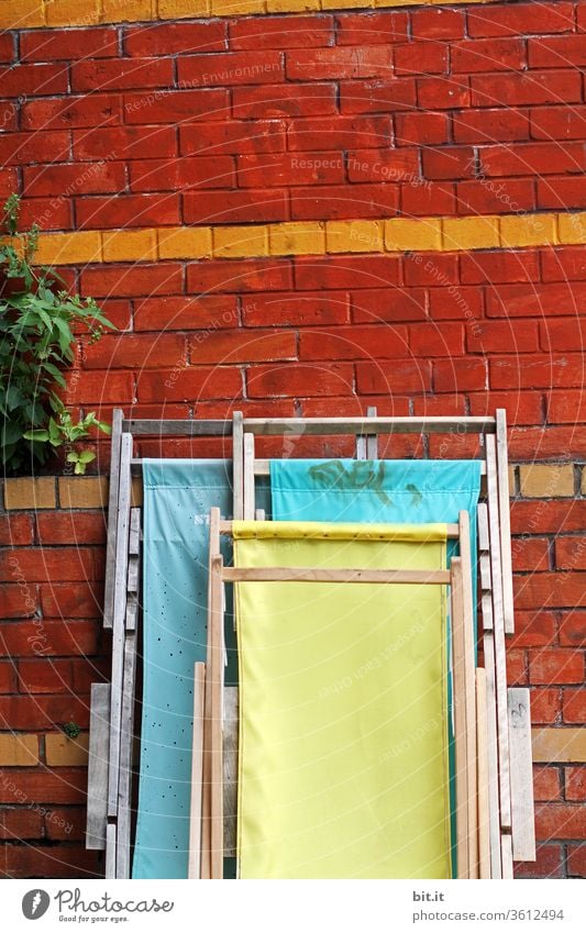 Drei einsame Liegestühle stehen zusammengeklappt, vor einer Wand aus Backstein und warten auf den Saisonanfang. Geschlossene, bunte Holzliegen lehnen ordentlich aufgeräumt an einer Wand mit Pflanze. Strandhaus im Sommer mit gelben & türkisen Stoffliegen.