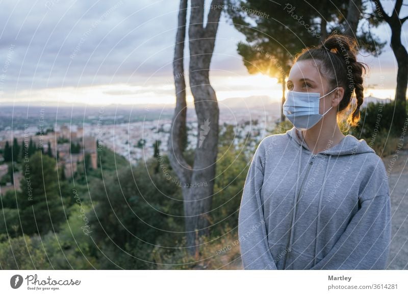 Junges Mädchen schaut mit einer Maske beim Covid, Verschmutzung durch die Sonne im Hintergrund, über der Stadt der Alhambra, Spanien. Sommerlicher Sonnenuntergang mit Sonnenlicht.