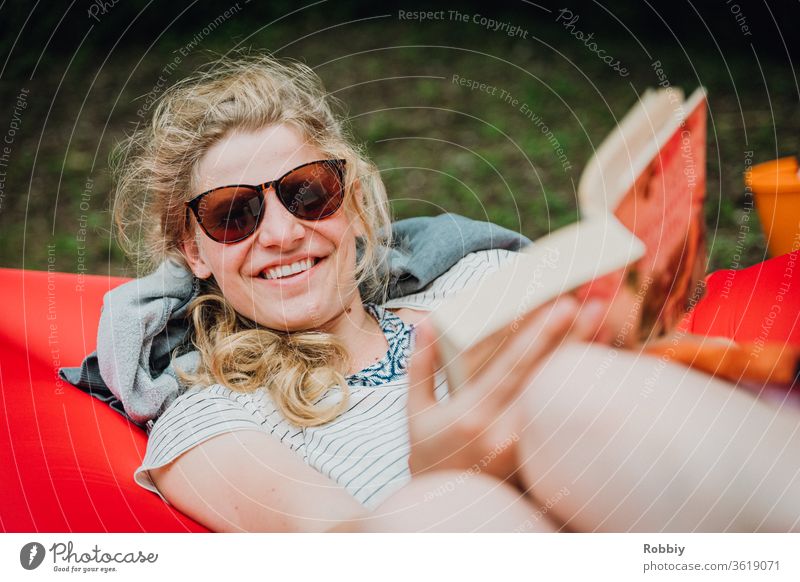 Junge Frau liest im Urlaub liegend auf einem Lamzac ein Buch und lächelt freundlich in die Kamera lesen Literatur lächeln Snnenbrille entspannen chillen relaxen