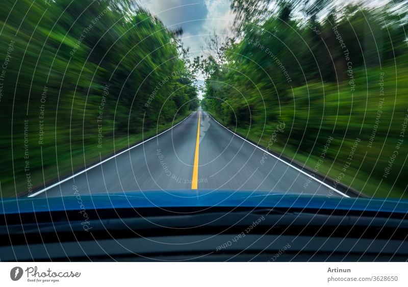 Frontansicht eines blauen Autos, das mit hoher Geschwindigkeit auf der Mitte einer Asphaltstraße mit weißer und gelber Linie des Verkehrssymbols im grünen Wald fährt. Die Bäume neben der Straße sind verschwommen. Auto mit verschwommener Bewegung.