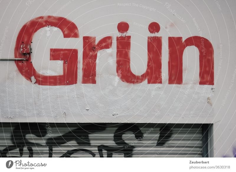 Schriftzug Grün in roten Buchstaben auf grauer Wand mit Jalousie, paradox oder widersprüchlich Widerspruch Paradox rot-grün farbenblind Mauer Fassade Gebäude