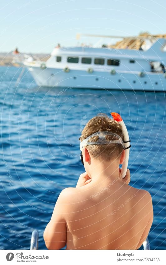 Ein Junge setzt eine Tauchermaske auf, bereit, im Meer zu schwimmen. Im Hintergrund ist eine weiße Yacht zu sehen. aktiv Strand blau Kind Konzept Korallen