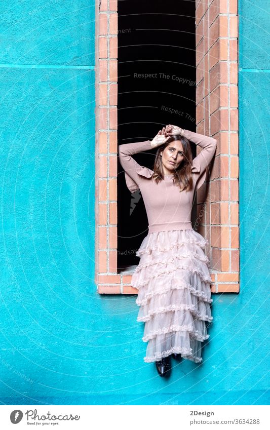 Mittelgroße, schöne Frau mit rosa Kleid, die an einem Fenster in einem modernen Gebäude sitzt 1 hübsch Porträt Mode Sitzen lange Haare Farben