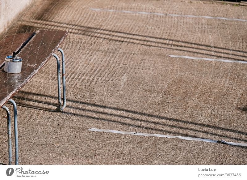 Ein Raucherplatz in der Sonne: braune Holzbank, Sternchen auf dem Aschenbecher, viel Sonne und lange Schatten am Boden Sitzplatz alt sonnig Sommer Linien einsam