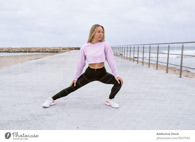 Fitnesssportlerin beim Ausfallschritt an der Böschung nahe des Ozeans Sportlerin Dehnung Bein Training Sportkleidung Stauanlage Meer Zaun Seite beweglich