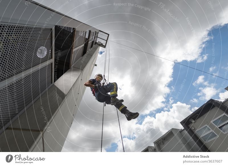 Feuerwehrmann klettert Seil gegen bewölkten Himmel herunter Training Aufstieg Gerät Mut stark Gebäude üben männlich retten komplex Uniform behüten Schutzhelm