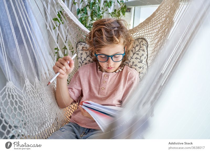 Brillenschüler lernt in Hängematte mit Heft zu Hause Schuljunge lernen Bildung Hausaufgabe Wissen intelligent fokussiert Schönschreibheft Bleistift Junge