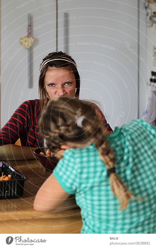 Zwei Teenager sitzen zuhause in der Küche am Tisch und schneiden Gemüse und Grimasse. Lustige Schwestern helfen daheim bei Hausarbeit, machen Quatsch und haben Spaß. Weibliche Geschwister von vorne und hinten, mit hübschen Frisuren. Pädagogisch wertvoll.