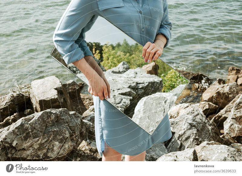 Nahaufnahme einer am Ufer stehenden Frau in blauem Kleid, die einen Spiegel hält, in dem sich Bäume und Felsen spiegeln, selektiver Fokus Erwachsener Sauberkeit