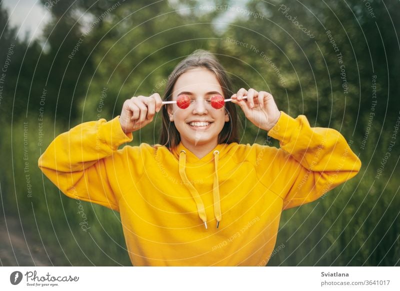 Ein lächelndes Mädchen in einem knallgelben Pullover hält einen knallroten Lolli in der Hand. Sommerliche Natur Bonbon Lollipop lustig hell Gesundheit Lifestyle