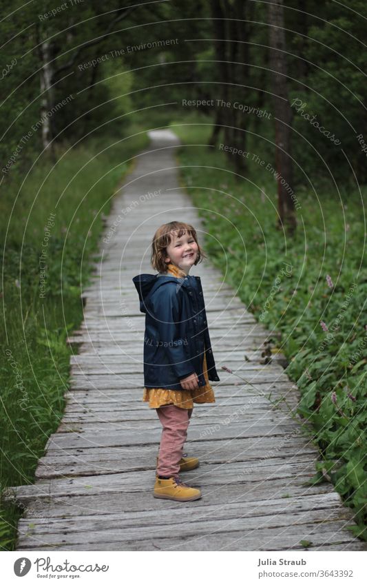 Mädchen mit Regenjacke freut sich, stehend auf einem Holzsteg in einem Wald Kleinkind süß stylisch gelb Knöterichgewächs Birkenwald Naturschutzgebiet Rhön