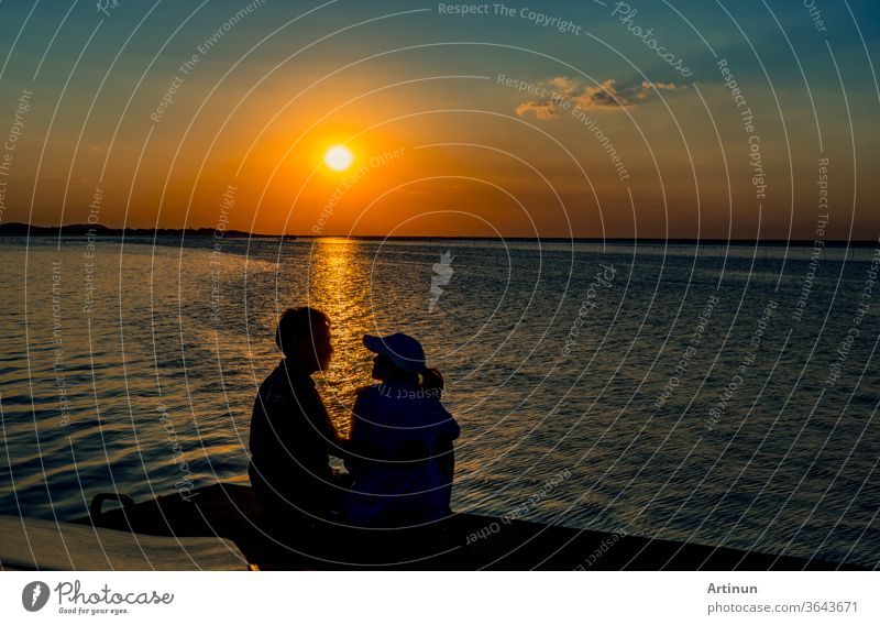 Silhouette eines verliebten Glückspaares, das bei Sonnenuntergang am Strand mit orangefarbenem und blauem Himmel sitzt und sich küsst. Sommerurlaub und Reisekonzept. Romantisches junges Paar, das sich am Meer trifft.