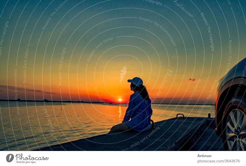 Einsame junge Frau trägt eine Mütze, die sich bei Sonnenuntergang allein vor dem Auto mit orangefarbenem und blauem Himmel am Strand entspannt. Sommerurlaub und Reisekonzept.