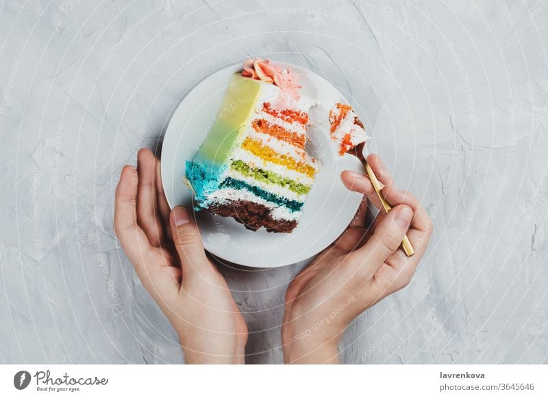 Flachgelegtes Regenbogen-Veganer-Kuchenstück in Frauenhänden auf grauem Hintergrund Diät Geschenk rosa Snack geschmackvoll Glück Mädchen Gebäck Hand Geburtstag