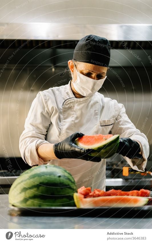 Köchin in Atemschutzgerät schneidet reife Wassermelone in Restaurantküche Frau Küchenchef Scheibe Fokus Uniform Konzentration Handschuh frisch Koch organisch