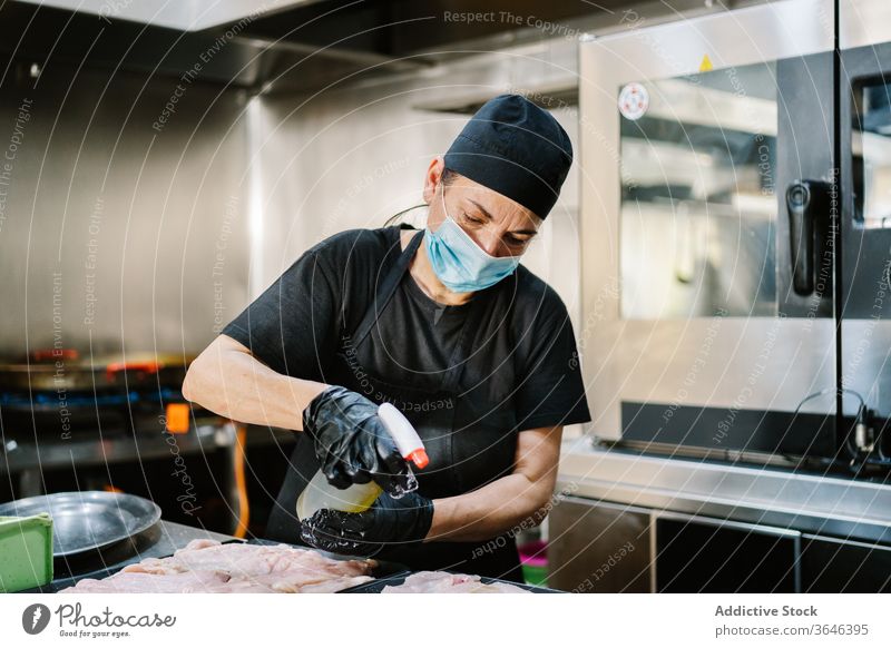 Fokussierte Köchin in Atemschutzgerät, die Fleisch auf Backblech sprüht Küchenchef Frau Spray Erdöl Hähnchen Pfanne Konzentration Coronavirus ernst Koch