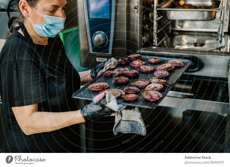 Getreidekoch in Atemschutzgerät mit gerösteter Zwiebel auf Backblech Küchenchef Frau Braten Ofen Fokus Restaurant Coronavirus Bestandteil Lebensmittel Koch