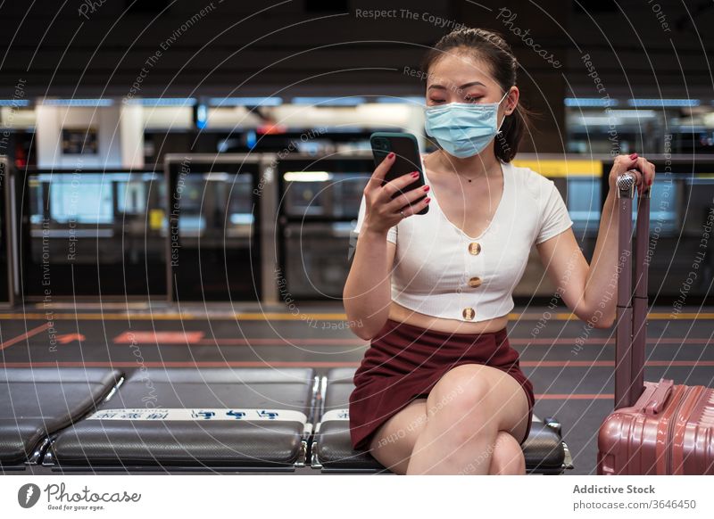 Asiatische Frau in Maske benutzt Smartphone in Flughafen-Zugverbindung benutzend Koffer Station Bahnverbindung reisen Mundschutz Atemschutzgerät Coronavirus