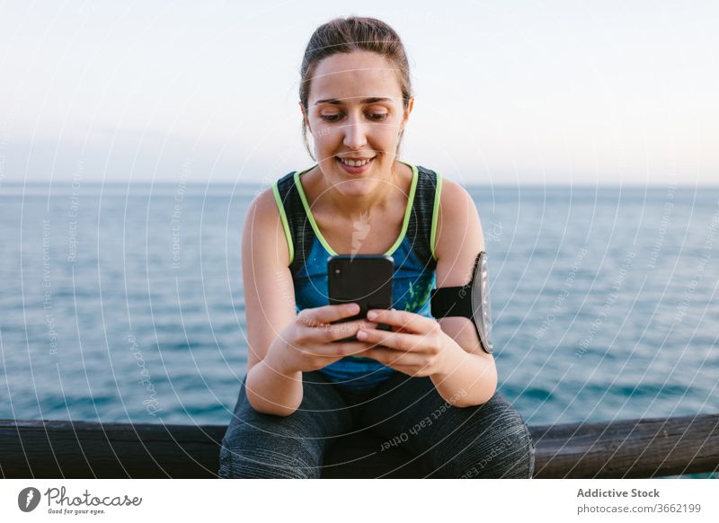 Lächelnde Frau in Sportkleidung mit Smartphone am Meer Sportlerin Stauanlage Browsen sich[Akk] entspannen Training MEER Sportbekleidung benutzend Fitness