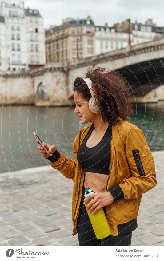 Sportlerin mit Headset nutzt Social Media auf Smartphone nach dem Training Kopfhörer soziale Netzwerke plaudernd Internet Pause Stauanlage Fluss benutzend