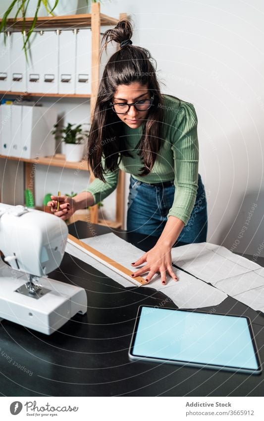 Ethnische Schneiderin mit Lineal in der Nähe der Nähmaschine in der Werkstatt Näherin messen Handwerk nach vorne lehnen Kleinunternehmen Arbeit Backsteinwand